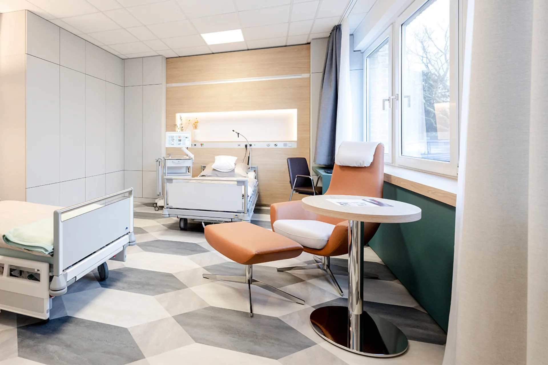 Modern eingerichtetes Patientenzimmer mit Krankenbett, Sessel, Hocker und rundem Beistelltisch vor Fenster