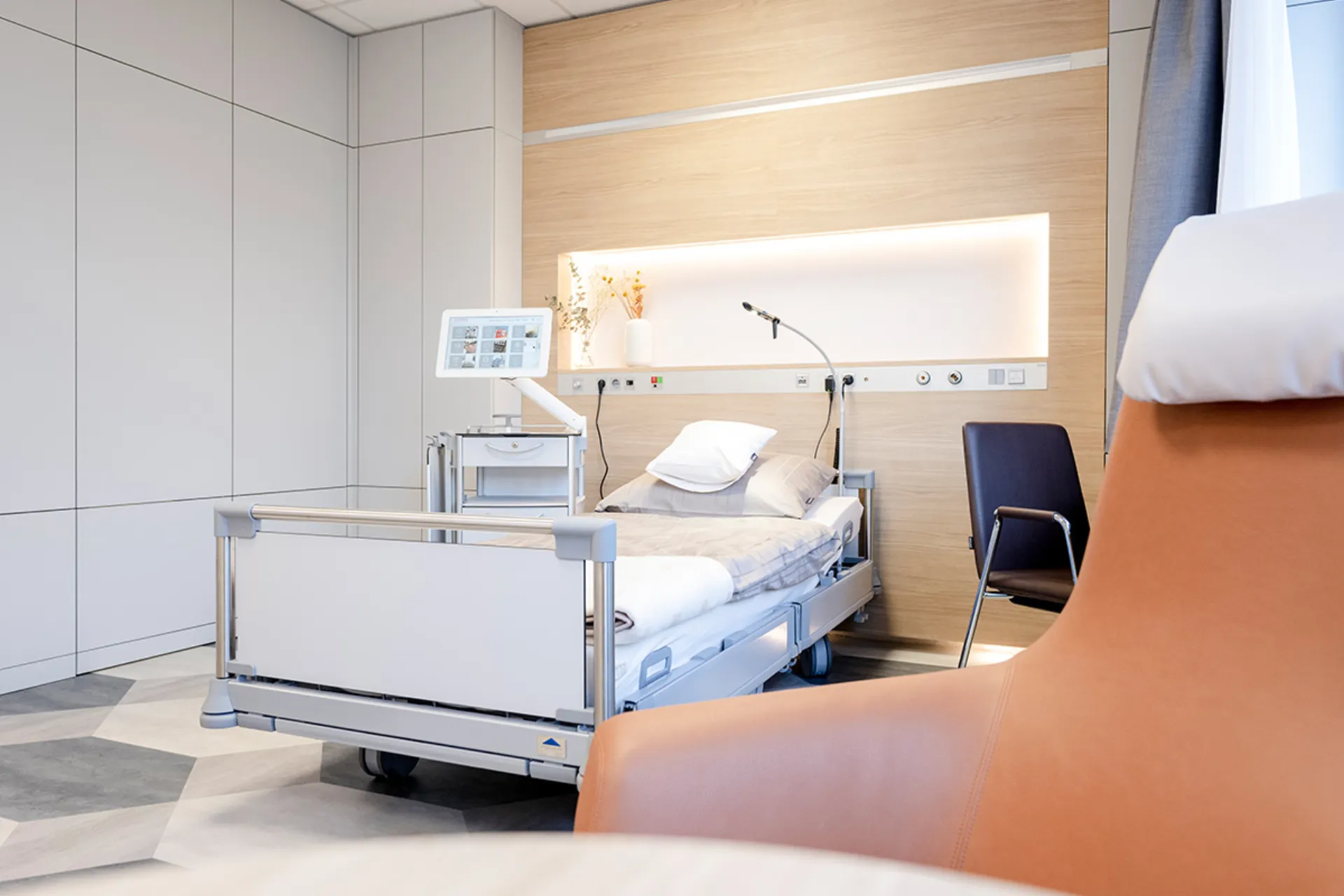 Krankenhauszimmer mit verstellbarem Bett, Leselampe, Monitor und bequemen Stuhl vor Fenster mit Vorhang