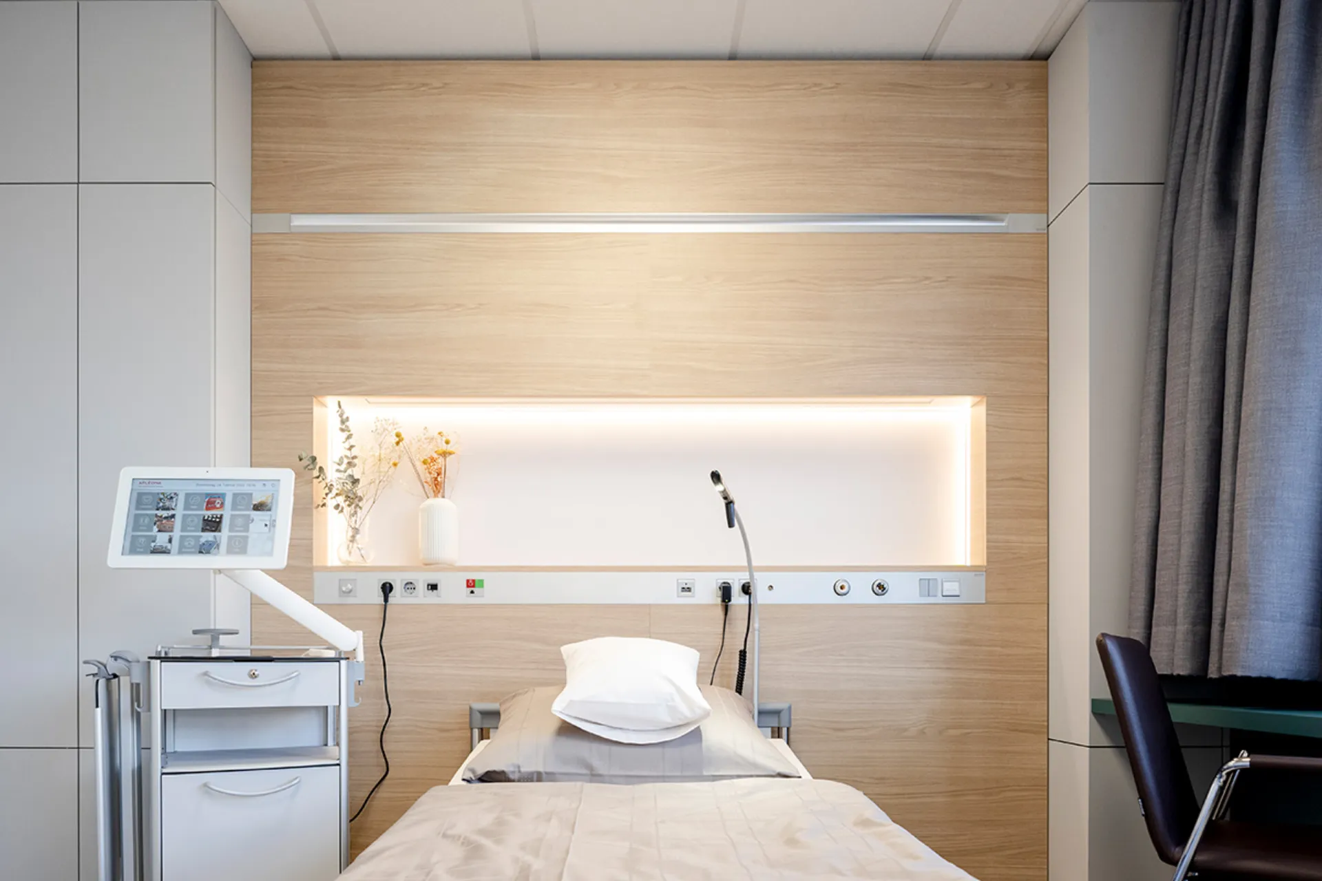 Krankenhauszimmer mit Einzelbett, Kopfkissen, medizinischem Monitor auf Rollwagen und Holzpaneelwand mit Beleuchtung und medizinischen Anschlüssen