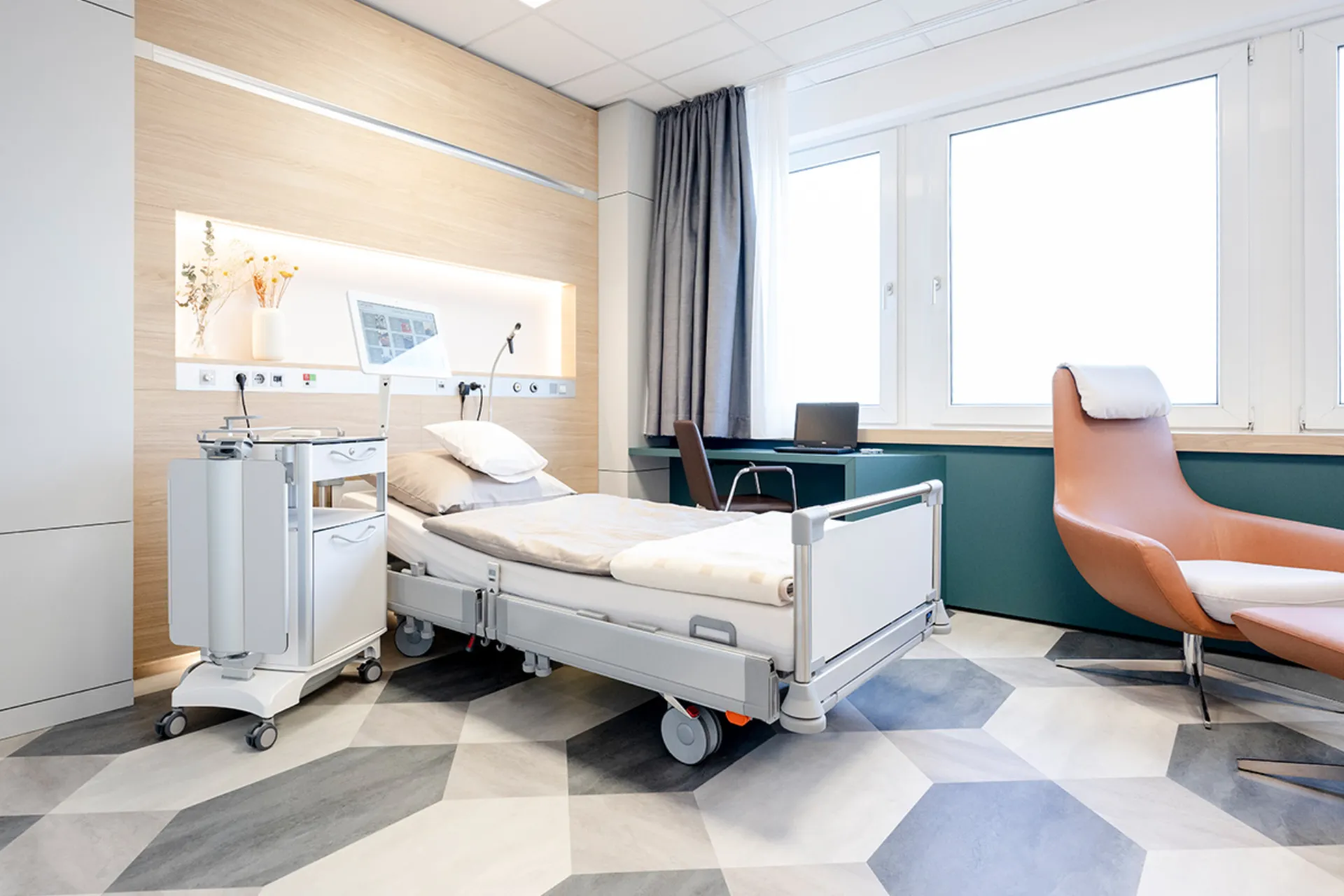 Modern eingerichtetes Patientenzimmer mit Pflegebett, medizinischem Gerät auf Rollen, Schreibtisch mit Stuhl und einem Sessel vor großen Fenstern