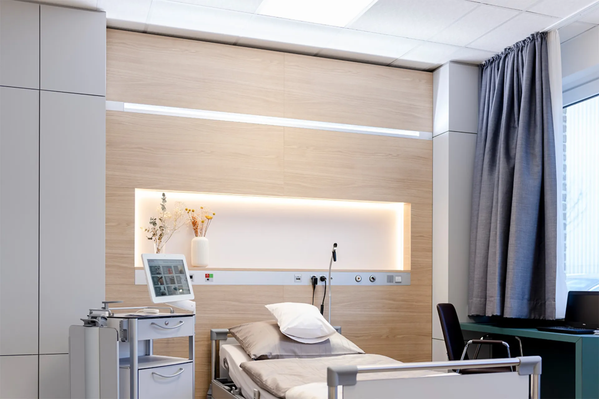 Krankenhauszimmer mit Einzelbett, medizinischem Gerät neben dem Bett, Holzverkleidung an der Wand und großem Fenster mit Vorhängen