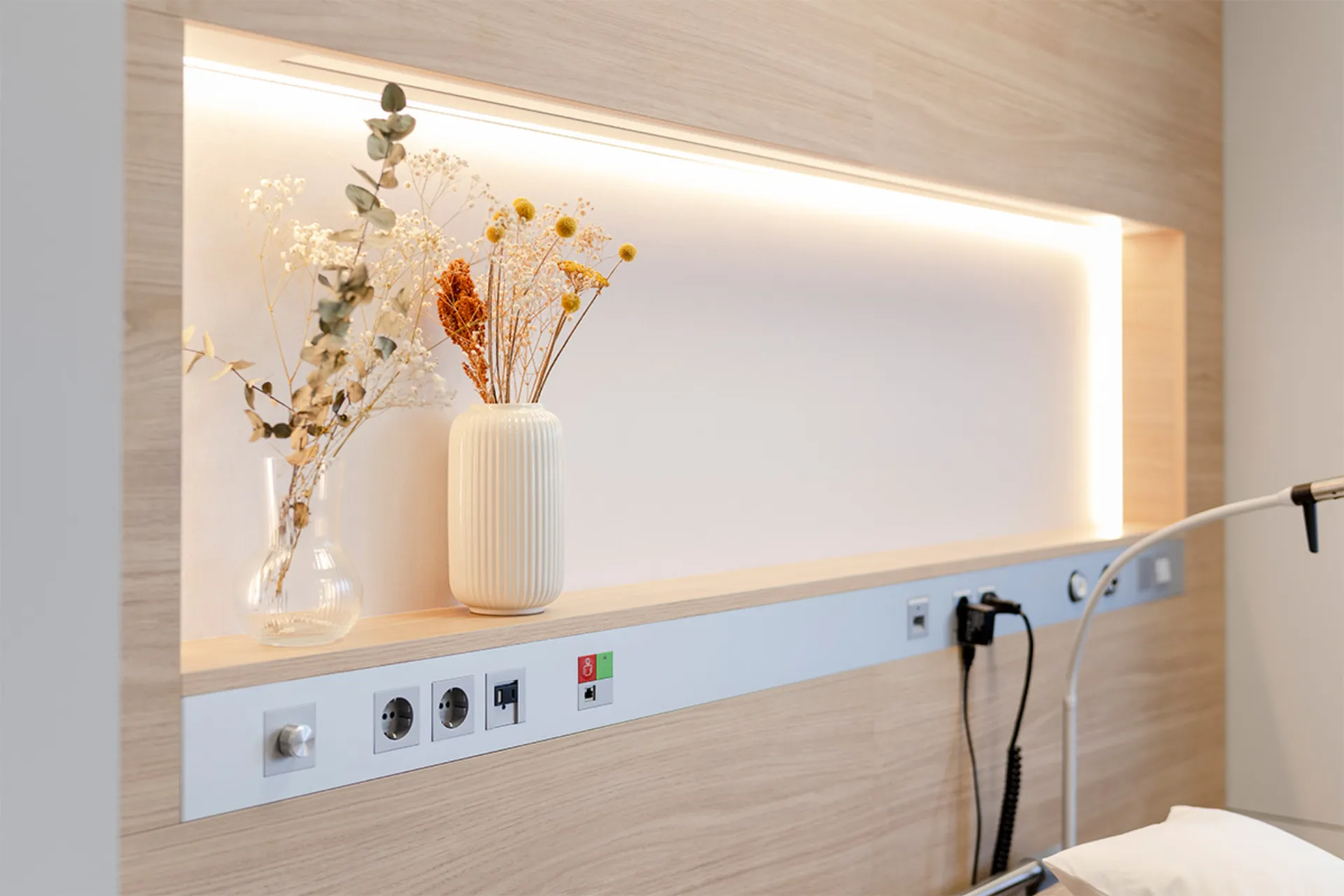 Nische mit indirekter Beleuchtung, zwei Vasen mit Trockenblumen und einer Reihe von Steckdosen an einer hellen Holzwand