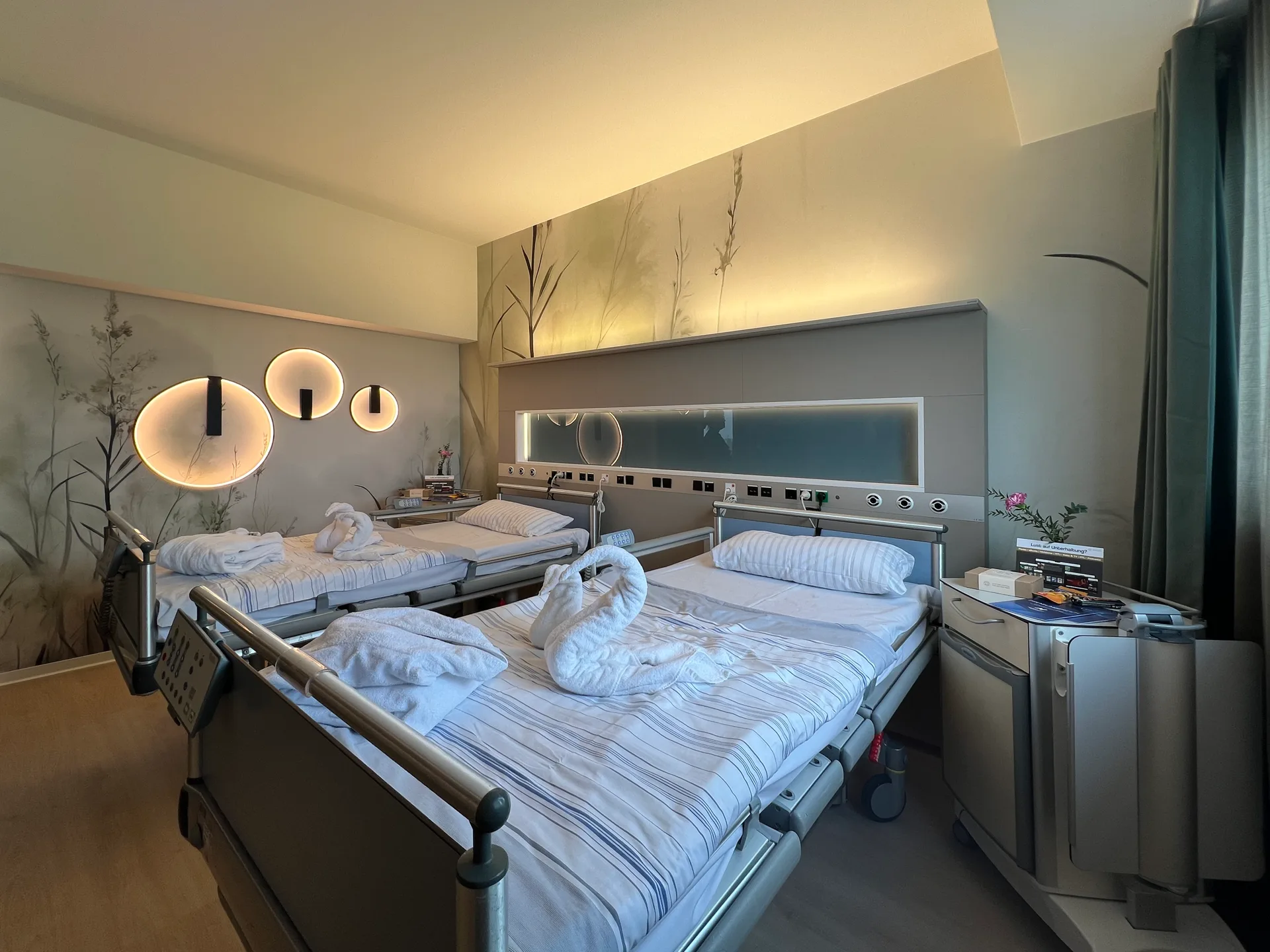 Zweibettzimmer mit Q100 integrativ vitra, indirekt beleuchtetem Wandpaneel mit Naturmotiv, runden Wandleuchten und Pflegebetten mit aus Handtüchern geformten Schwänen in warmer Abendbeleuchtung