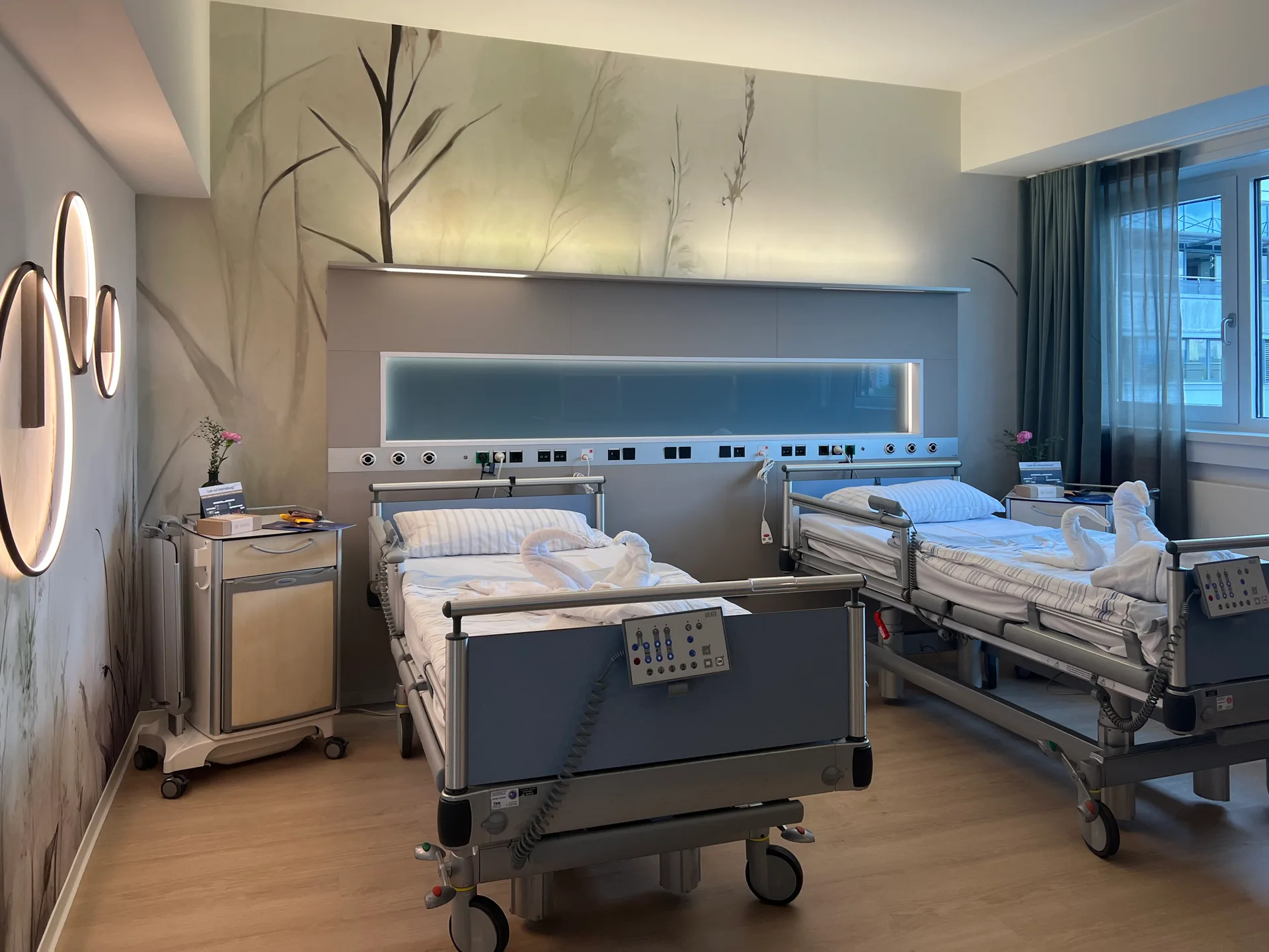 Zweibettzimmer mit Q100 integrativ vitra, hinterleuchtetem Wandpaneel mit Gräsermotiv, Pflegebetten mit Handtuchschwänen und Fenster mit Vorhängen bei Tageslicht