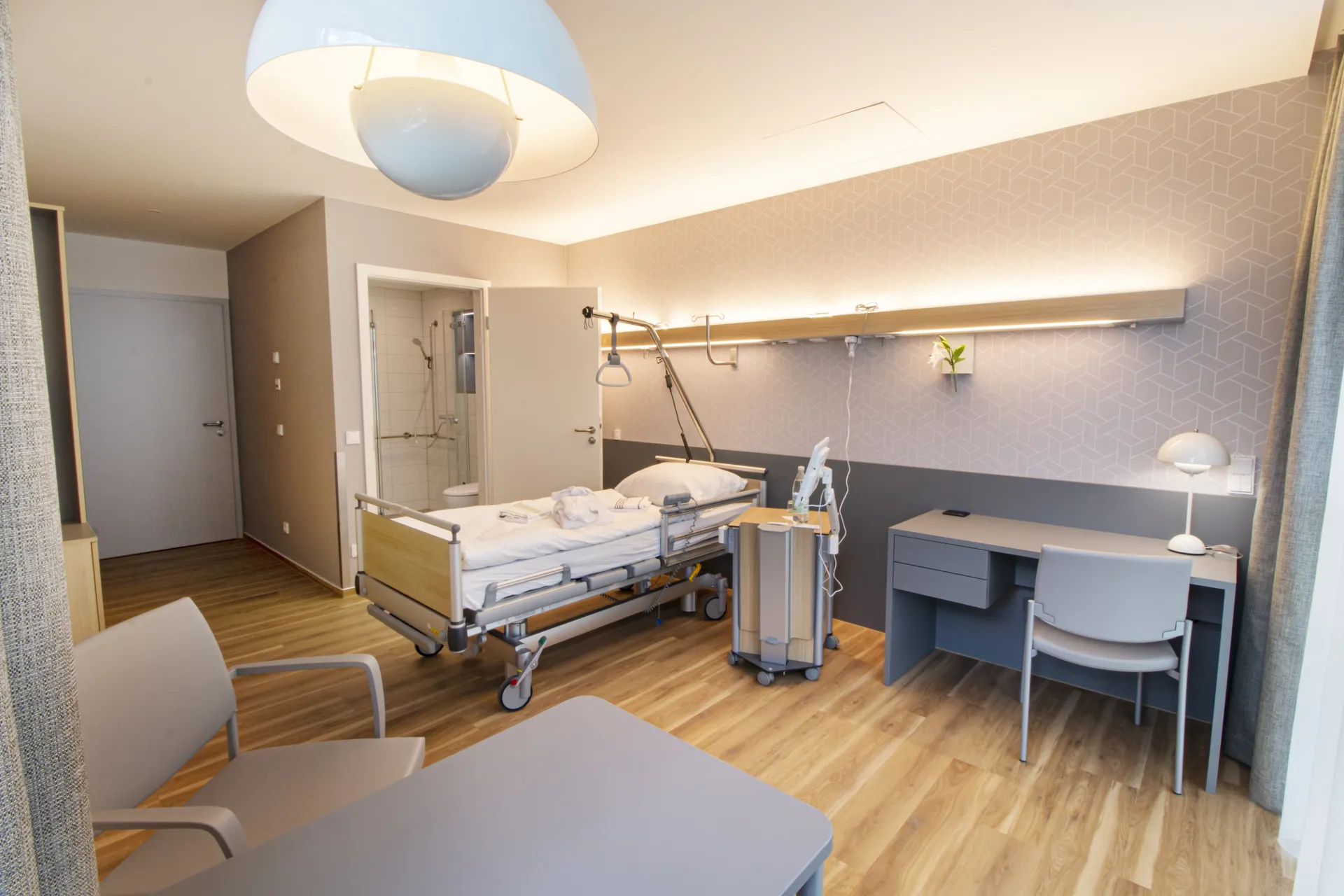 Modernly furnished patient room with hospital bed, desk, chairs and en-suite bathroom