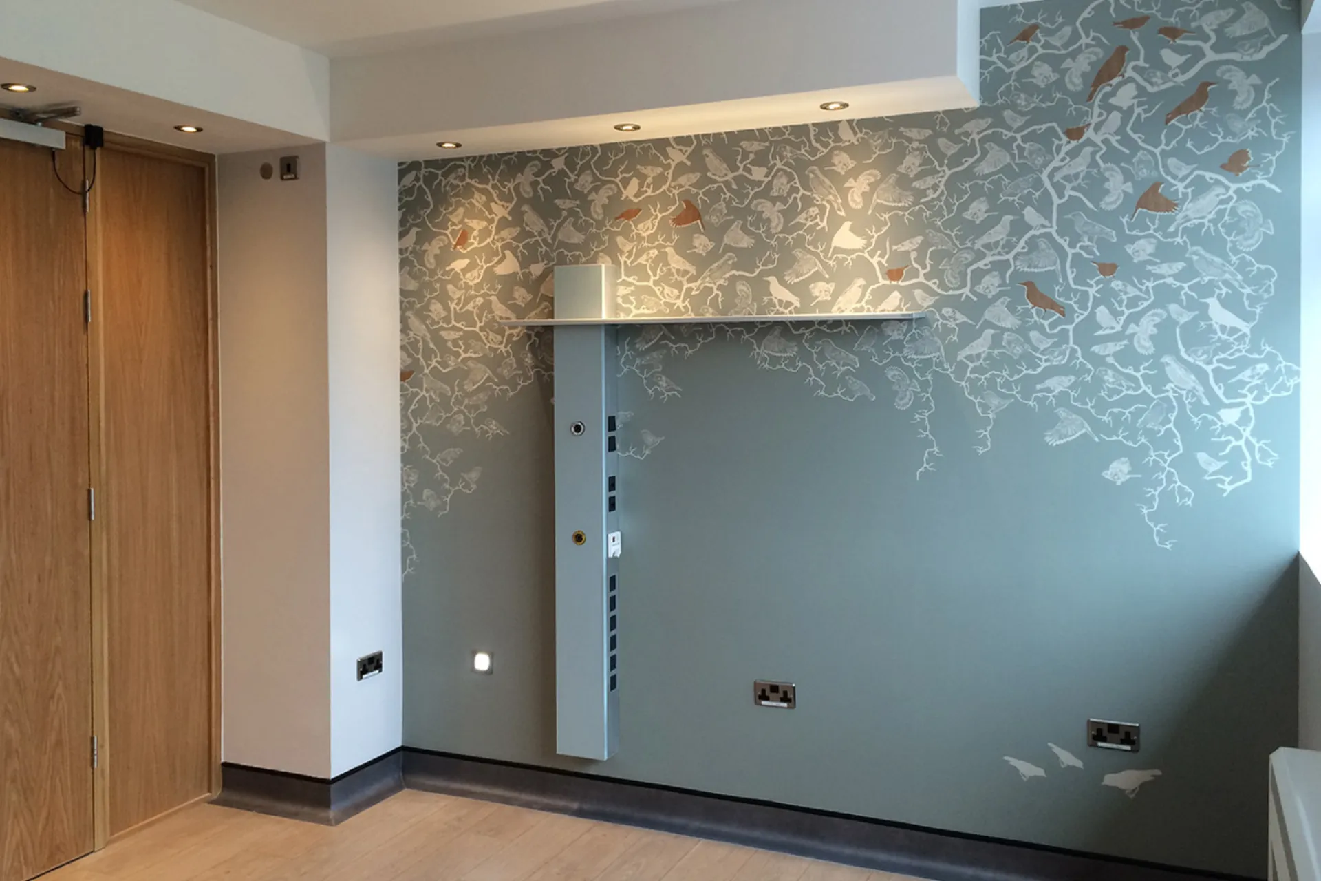 Wall with light-grey wallpaper and a white, branching pattern next to a wooden door and a white shelf