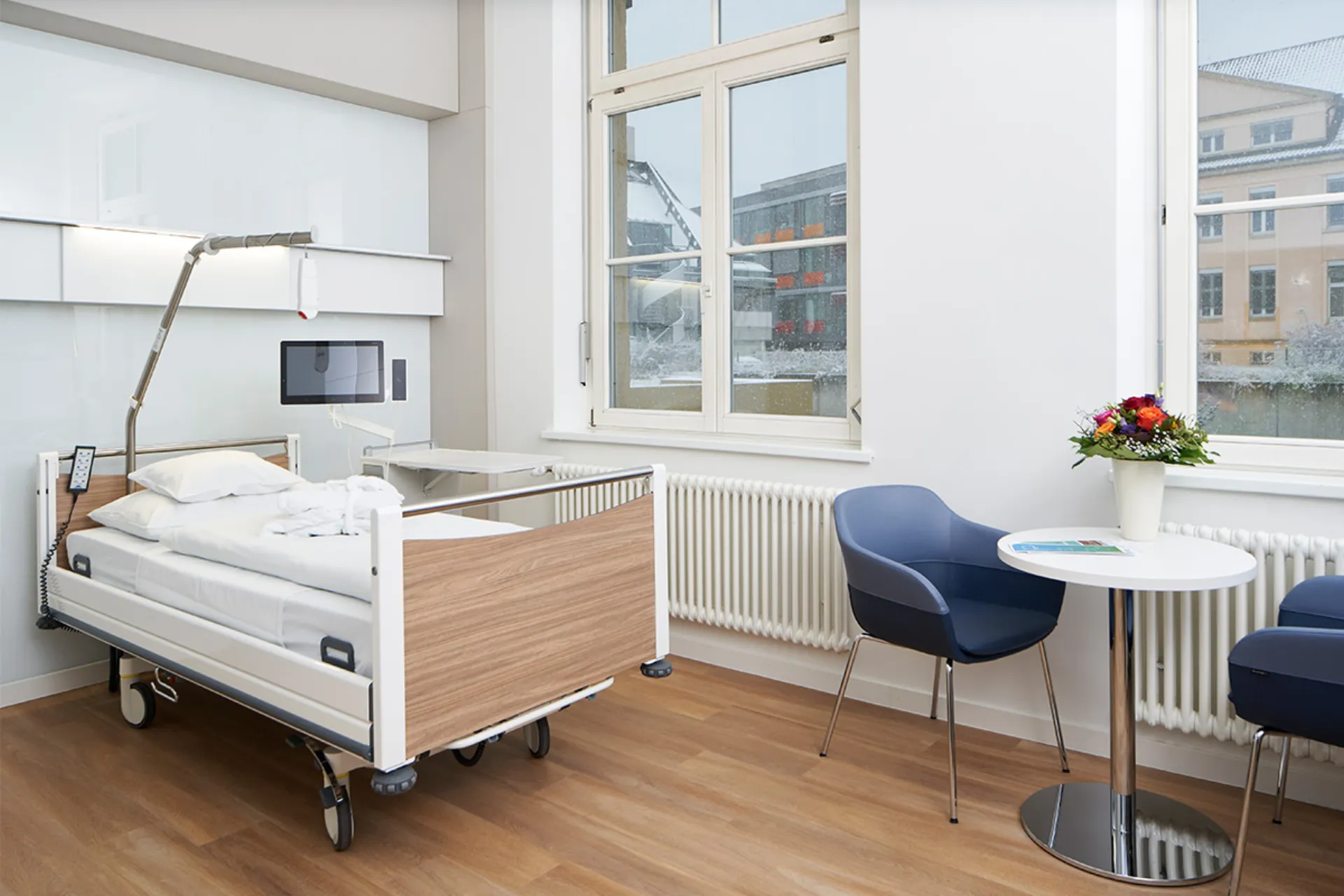 Hospital room with care bed, two blue chairs, a round table with a flower vase and large windows