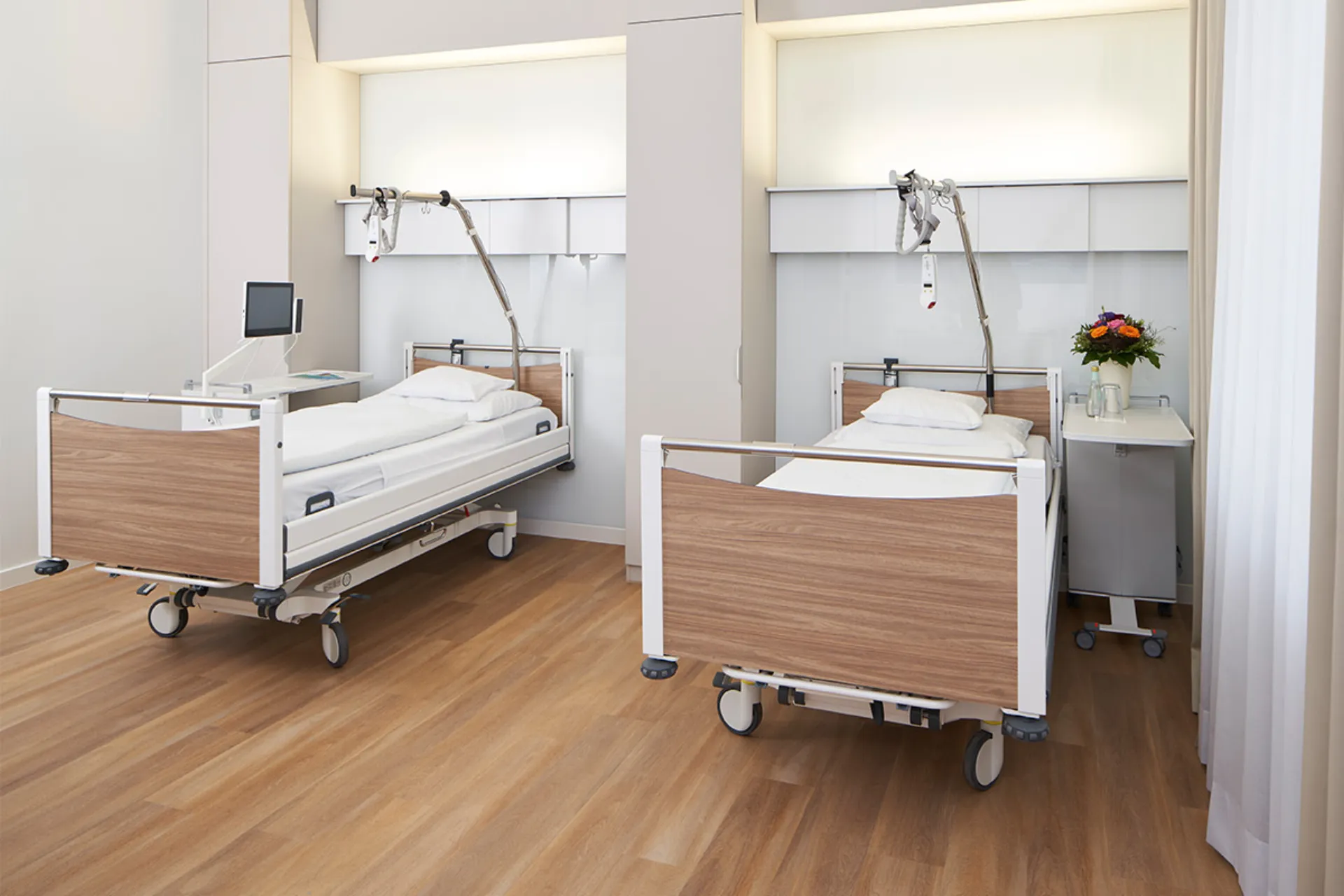Two modern hospital beds with wood panelling in a bright patient room with medical devices and a bouquet of flowers on a trolley table