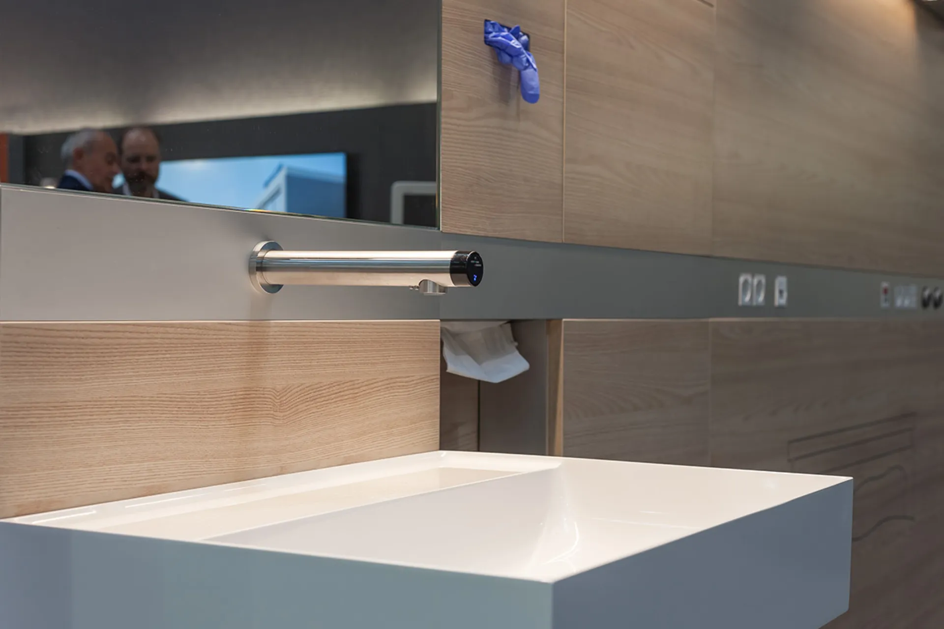 Modern washbasin with a rectangular bowl, a touchless tap and wood panelling on the wall, with a mirror and a blue glove holder