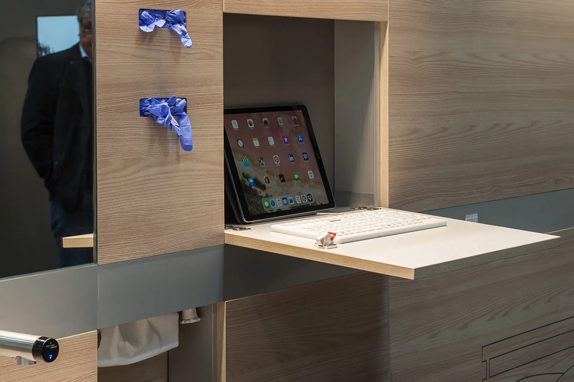 Tablet with numerous app icons standing on a pull-out wooden tray next to a white keyboard in a modern wooden shelf unit